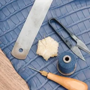 Leatherworking tools arranged on blue crocodile leather include a metal straightedge, thread snips, a pin cushion with needles, a spool of blue thread, and a wooden-handled awl.
